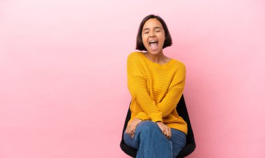 Genç melez kadın pembe arka planda izole edilmiş bir sandalyede oturuyor ve ağzı açık bir şekilde öne doğru bağırıyor.