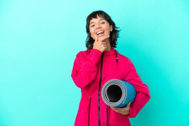 Mavi arka planda bir minder tutarken yoga derslerine giden genç kız mutlu ve gülümsüyor.