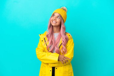 Pembe saçlı, yağmura dayanıklı bir ceket giyen genç bir kadın mavi arka planda izole edilmiş gülümserken yukarı bakıyor.