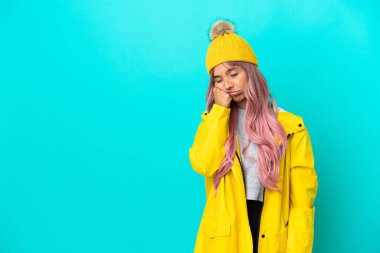 Pembe saçlı, yağmura dayanıklı bir ceket giyen, yorgun ve sıkılmış bir ifadeyle mavi arka planda izole edilmiş genç bir kadın.
