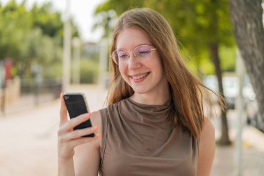 Gözlüklü Fransız kız dışarıda cep telefonu kullanıyor, mutlu bir ifadeyle.