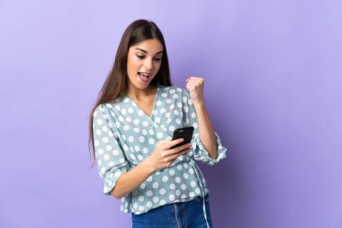 Genç beyaz kadın mavi arka planda izole edilmiş ve telefon zafer pozisyonunda.