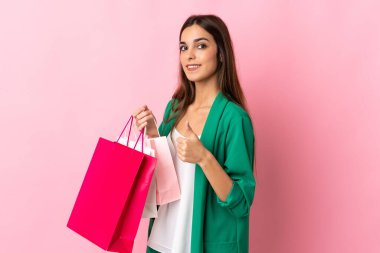 Pembe arka planda izole edilmiş genç beyaz kadın elinde alışveriş torbaları ve başparmağıyla