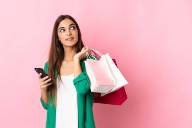 Pembe arka planda izole edilmiş beyaz bir kadın elinde alışveriş çantaları ve bir cep telefonu tutuyor.