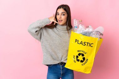 Pembe arka planda izole edilmiş plastik şişelerle dolu bir çanta taşıyan beyaz bir kadın telefon hareketi yapıyor. Bana geri dönüş işareti ver.