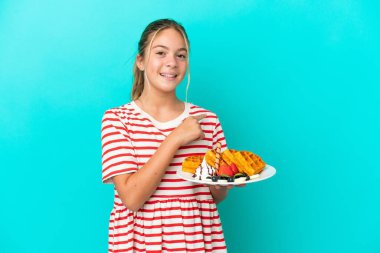 Mavi arka planda izole edilmiş waffle tutan küçük beyaz kız bir ürünü sunmak için yan tarafı işaret ediyor.