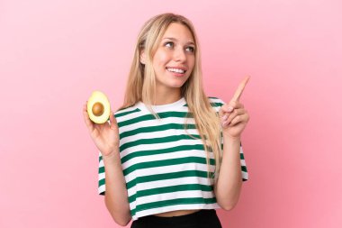 Pembe arka planda izole edilmiş avokado tutan beyaz bir kadın harika bir fikre işaret ediyor.