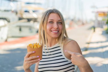 Sarışın genç bir kadın, açık havada yüzünde sürpriz bir ifadeyle patates kızartması tutuyor.