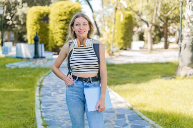 Açık havada genç sarışın bir kadın elinde mutlu bir ifadeyle bir defter tutuyor.