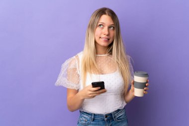 Mor arka planda izole edilmiş genç sarışın bir kadın elinde kahve ve bir şeyler düşünürken bir cep telefonu tutuyor.