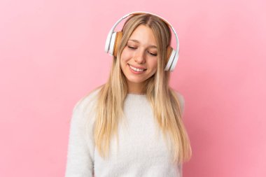 Pembe arka planda izole edilmiş genç sarışın kadın müzik dinliyor.