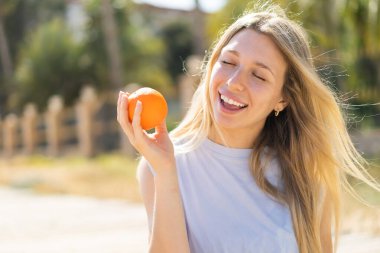 Açık havada genç sarışın bir kadın elinde mutlu bir ifadeyle bir portakal tutuyor.