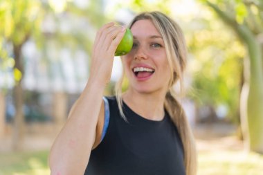 Açık havada genç sarışın bir kadın elinde elmayla şaşkın bir ifadeyle.