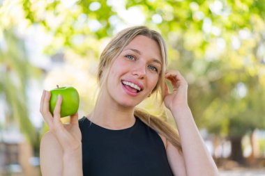 Açık havada elinde mutlu bir ifadeyle bir elma tutan genç sarışın kadın.