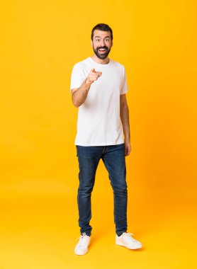 Sarı arka planda sakallı bir adamın tam boy fotoğrafı. Şaşırmış ve ön tarafı işaret ediyor.