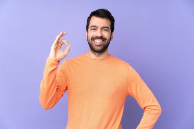 İzole edilmiş mor arka planda beyaz, yakışıklı bir adam parmaklarıyla tamam işareti gösteriyor.