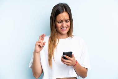 Mavi arka planda izole edilmiş genç beyaz kadın parmakları çapraz cep telefonu kullanıyor.