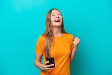 Genç Rus kadın mavi arka planda izole edilmiş ve telefon zafer pozisyonunda.