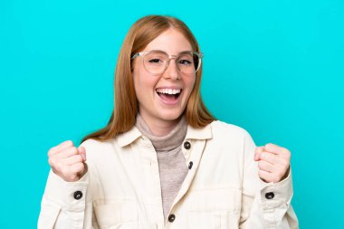 Mavi arka planda, gözlüklü ve zaferi kutlayan genç kızıl saçlı kadın.