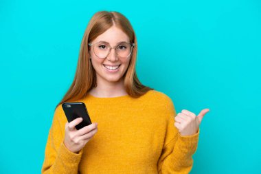 Genç kızıl saçlı kadın mavi arka planda izole edilmiş cep telefonu kullanıyor ve yan tarafı işaret ediyor.