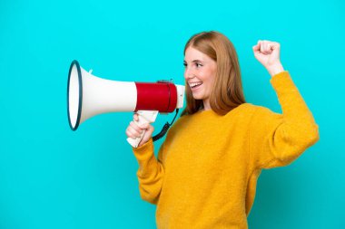 Genç kızıl saçlı kadın mavi arka planda izole edilmiş bir şekilde megafonla bir şey duyurmak için bağırıyor.