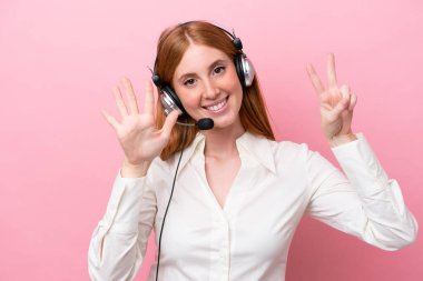 Pembe arka planda izole edilmiş kulaklıkla çalışan pazarlamacı kızıl saçlı kadın yediyi parmaklarıyla sayıyor.