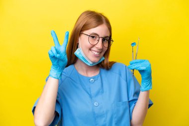 Genç kızıl saçlı dişçi kadın sarı arka planda izole edilmiş gülümsüyor ve zafer işareti gösteriyor.