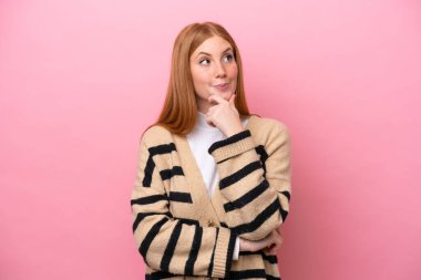 Genç kızıl saçlı kadın pembe arka planda izole edilmiş bir şekilde bakarken bir fikir düşünüyor.