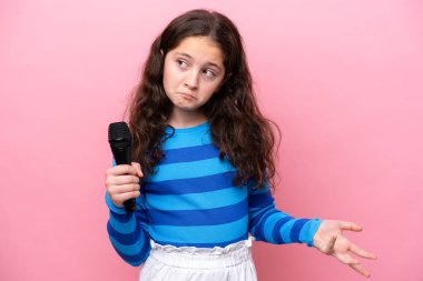 Küçük şarkıcı kız pembe arka planda izole edilmiş bir mikrofonu kaldırırken omuzlarını kaldırırken şüphe uyandırıyor.