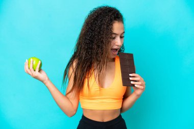 Genç bir Arap kadın mavi arka planda izole edilmiş. Bir elinde çikolata tableti, diğerinde elma alıyor.