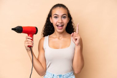 Elinde bej arka planda izole edilmiş saç kurutma makinesi tutan genç bir kadın harika bir fikre işaret ediyor.