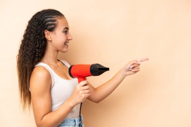 Elinde bej arka planda izole edilmiş saç kurutma makinesi olan genç bir kadın bir ürünü sunmak için kenarı işaret ediyor.