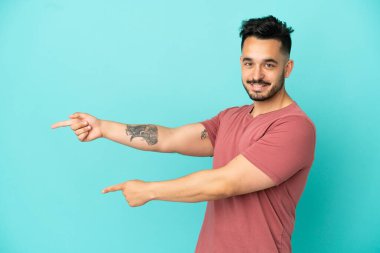 Mavi arka planda izole edilmiş genç beyaz bir adam parmağını yana doğru uzattı ve bir ürün sundu.