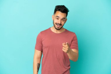 Mavi arka planda izole edilmiş genç beyaz adam şaşırmış ve ön tarafı işaret ediyor.