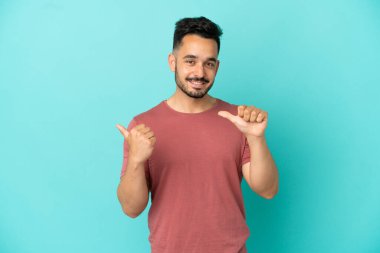 Mavi arka planda izole edilmiş genç beyaz adam bir ürünü sunmak için kenarı işaret ediyor.