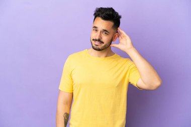 Mor arka planda izole edilmiş genç beyaz adam bir şey dinlerken elini kulağına koydu.