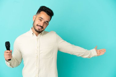 Beyaz tenli genç şarkıcı, mavi arka planda izole edilmiş. Gelmesi için ellerini yana uzatıyor.