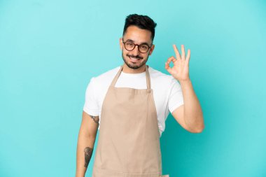 Restoran garsonu Kafkasyalı adam mavi arka planda izole edilmiş. Parmaklarıyla tamam işareti gösteriyor.