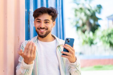 Yakışıklı bir Arap açık havada cep telefonu kullanıyor. Eli ile gelmesi için davet ediyor. Gelmene sevindim.