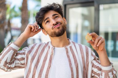 Yakışıklı bir Arap açık havada Bitcoin tutuyor kuşkuları var ve kafası karışmış bir ifadeyle.