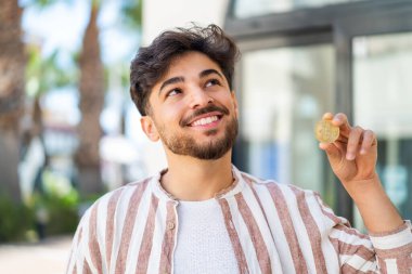 Yakışıklı bir Arap, Bitcoin 'i elinde tutuyor ve gülümserken yukarı bakıyor.