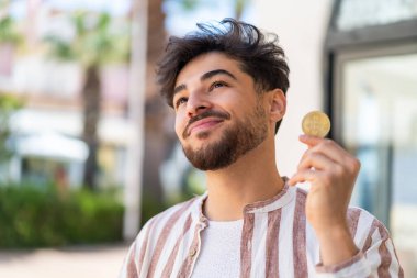 Açık havada elinde Bitcoin tutan yakışıklı bir Arap.