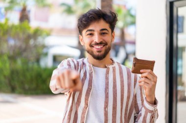 Açık havada cüzdan tutan yakışıklı bir Arap, kendine güvenen bir ifadeyle seni işaret ediyor.