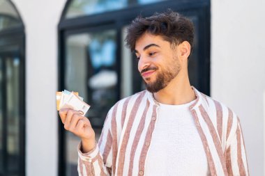 Dışarıdaki yakışıklı Arap adam mutlu bir ifadeyle bir sürü para alıyor.