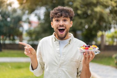 Genç Arap yakışıklı bir adam açık havada bir kase meyve tutuyor. Şaşırmış ve parmağıyla işaret ediyor.