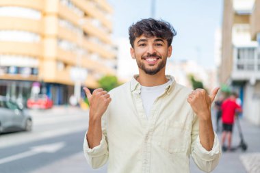Açık havada genç Arap yakışıklı bir adam. Baş parmakları havada ve gülümsüyor.