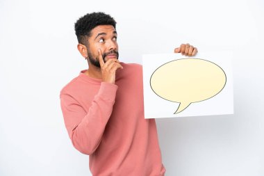 Genç Brezilyalı bir adam beyaz arka planda izole edilmiş. Elinde konuşma balonu ikonu olan bir levha var ve düşünüyor.