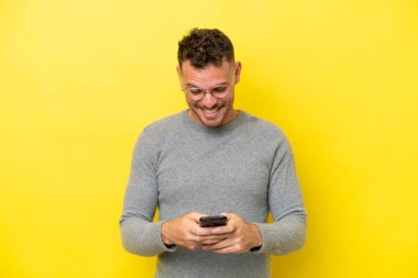 Sarı arka planda izole genç beyaz adam cep telefonuyla bir mesaj gönderiyor.