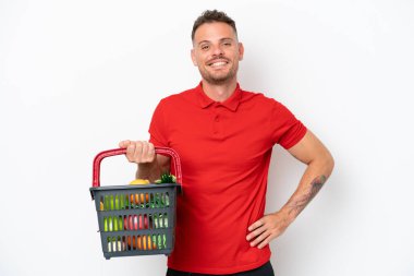 Beyaz arka planda izole edilmiş, kolları kalçasında gülümseyerek poz veren yiyecek dolu bir alışveriş sepeti tutan genç beyaz bir adam.