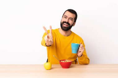 Beyaz bir adam kahvaltı yapıyor, gülümsüyor ve zafer işareti gösteriyor..